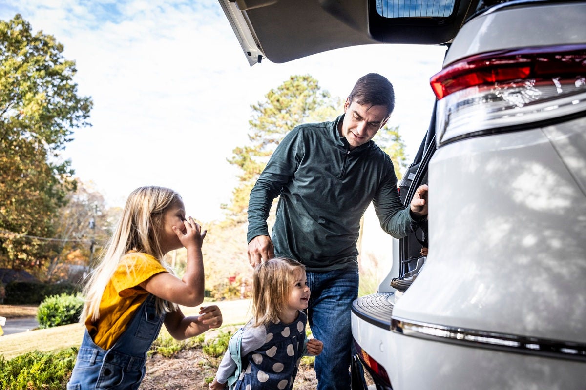 A person loads items into the back of a vehicle while two young children stand nearby outdoors.
