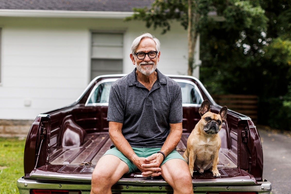 A person sits on the tailgate of a classic truck with a small dog seated beside them in a yard.