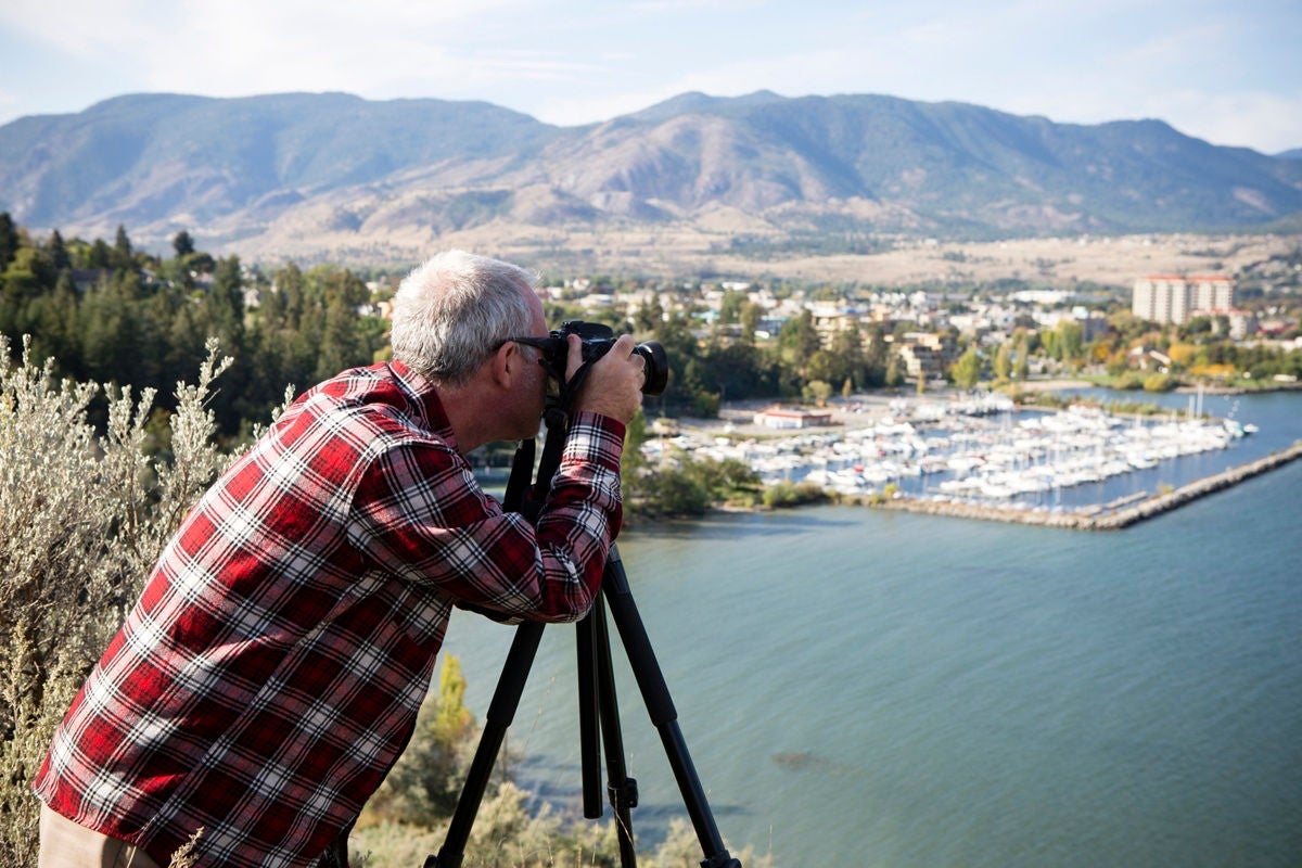 A person photographs a lakeside marina and mountain landscape using a camera on a tripod.