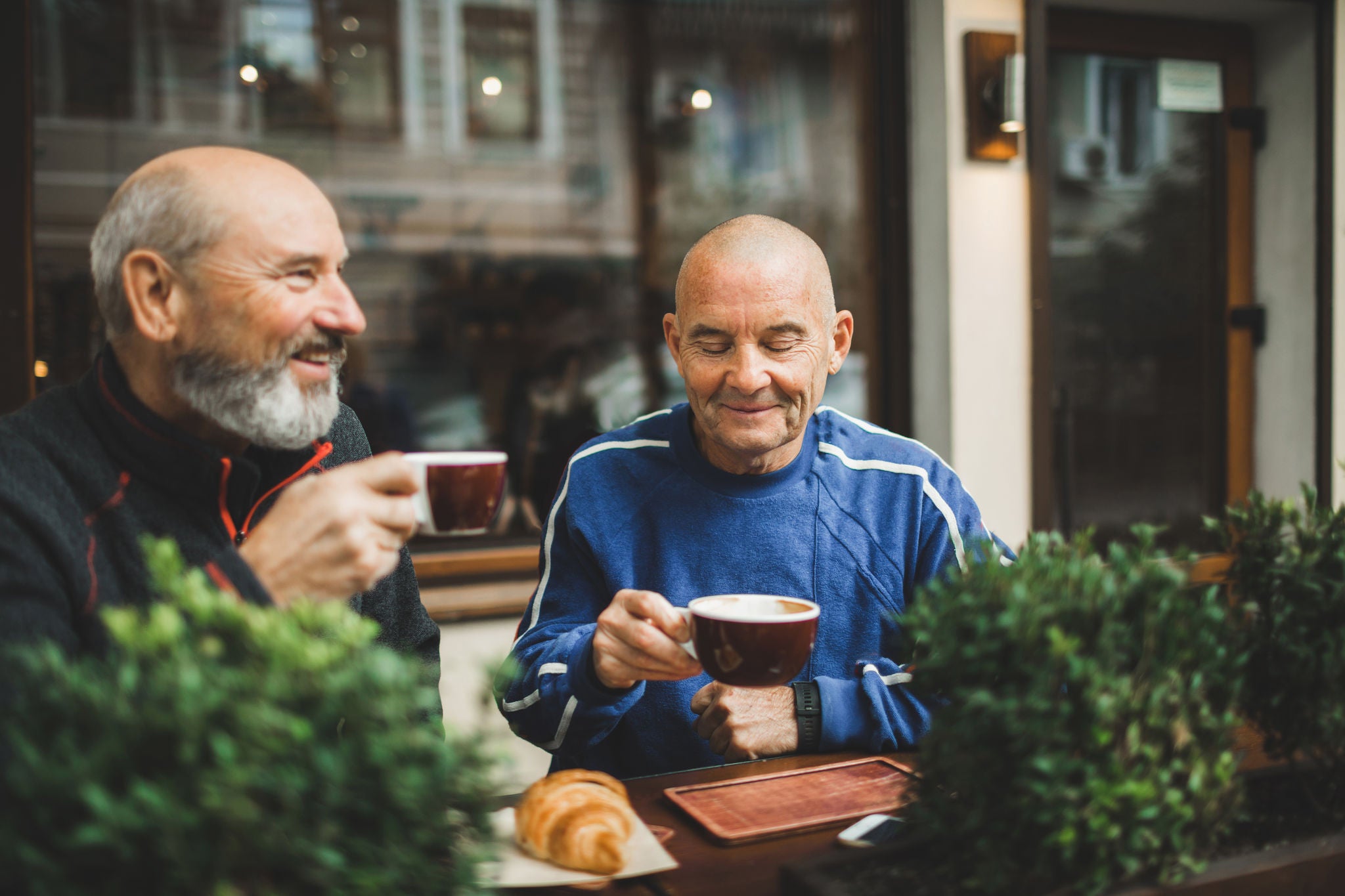 Two old age senior friends drinking coffee in cafe on outside terrace. Laughing and talking. Young at heart and active seniors. One with grey-haired beard. Positive emotions and lifestyle of retired people. Pavement cafe, european culture. Morning in city. Friendship concept.