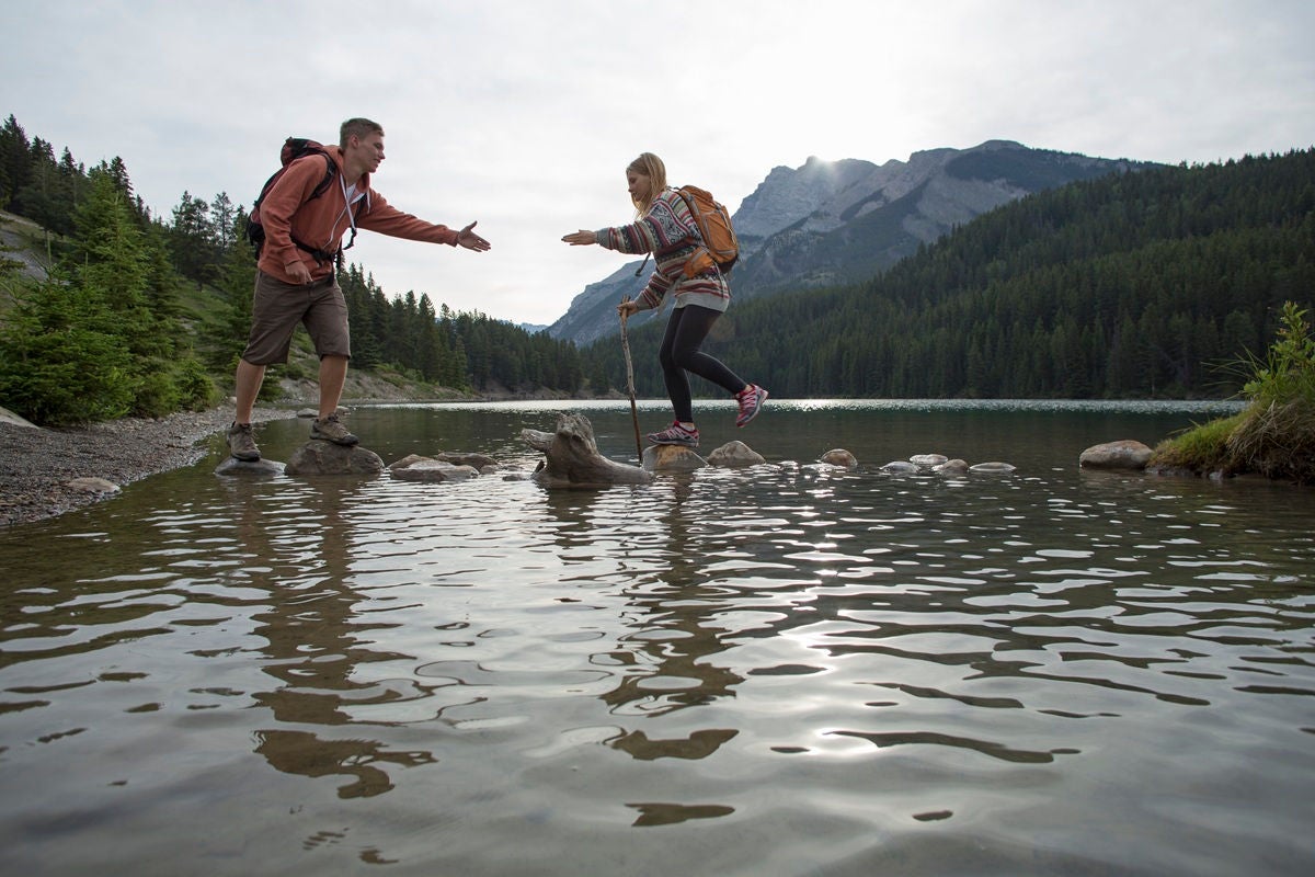 A person reaches out to help another person stepping across rocks in a calm mountain lake, with forested hills and peaks in the background.