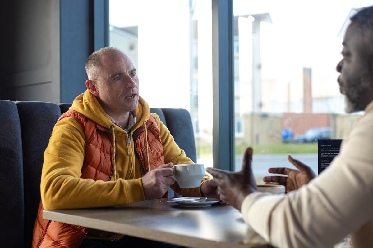 Two people seated at a café table by a window, holding coffee cups and talking together in a bright, casual setting.