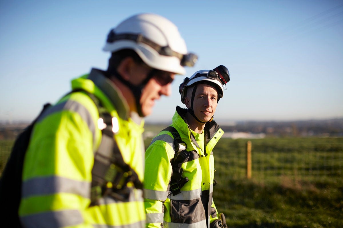 Two engineers working in a wind farm in high‑visibility gear and helmets stand outdoors at a worksite, talking under bright daylight.