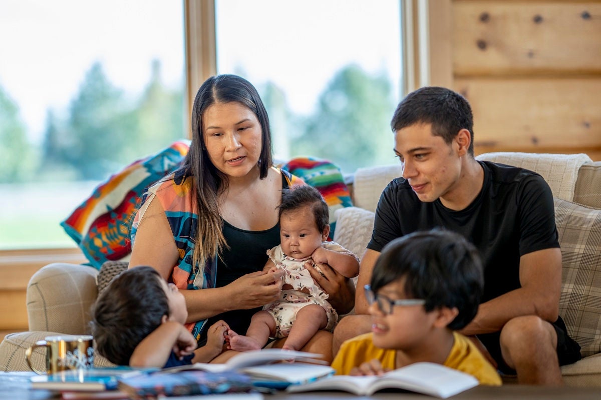 A family sits together on a couch, with adults holding a baby while two young children look on with books nearby.