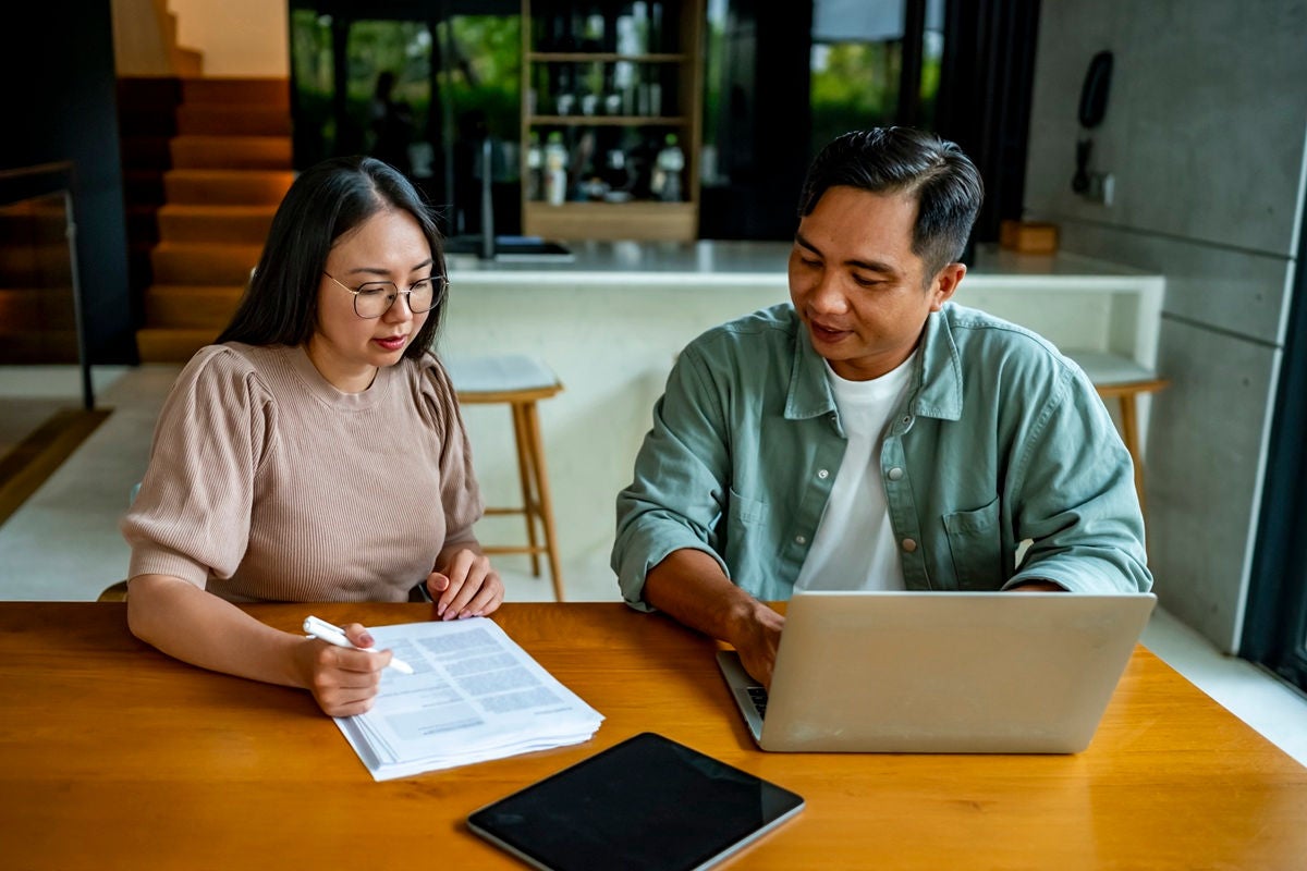 Two people sit at a table reviewing documents, with one using a laptop and a tablet resting nearby.