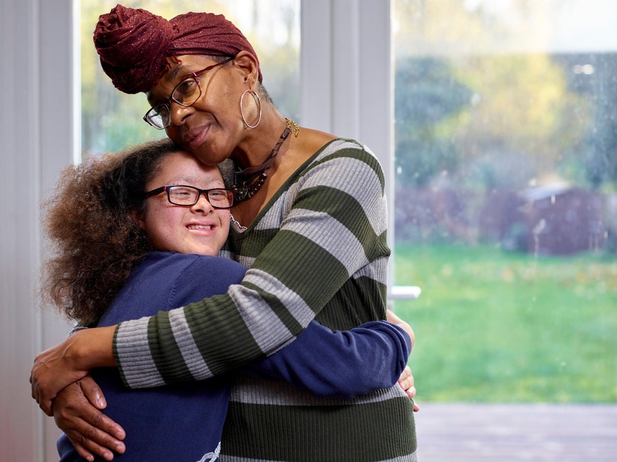 Adult and child standing indoors by a window, sharing a close embrace in a softly lit room with a view of a green backyard outside.