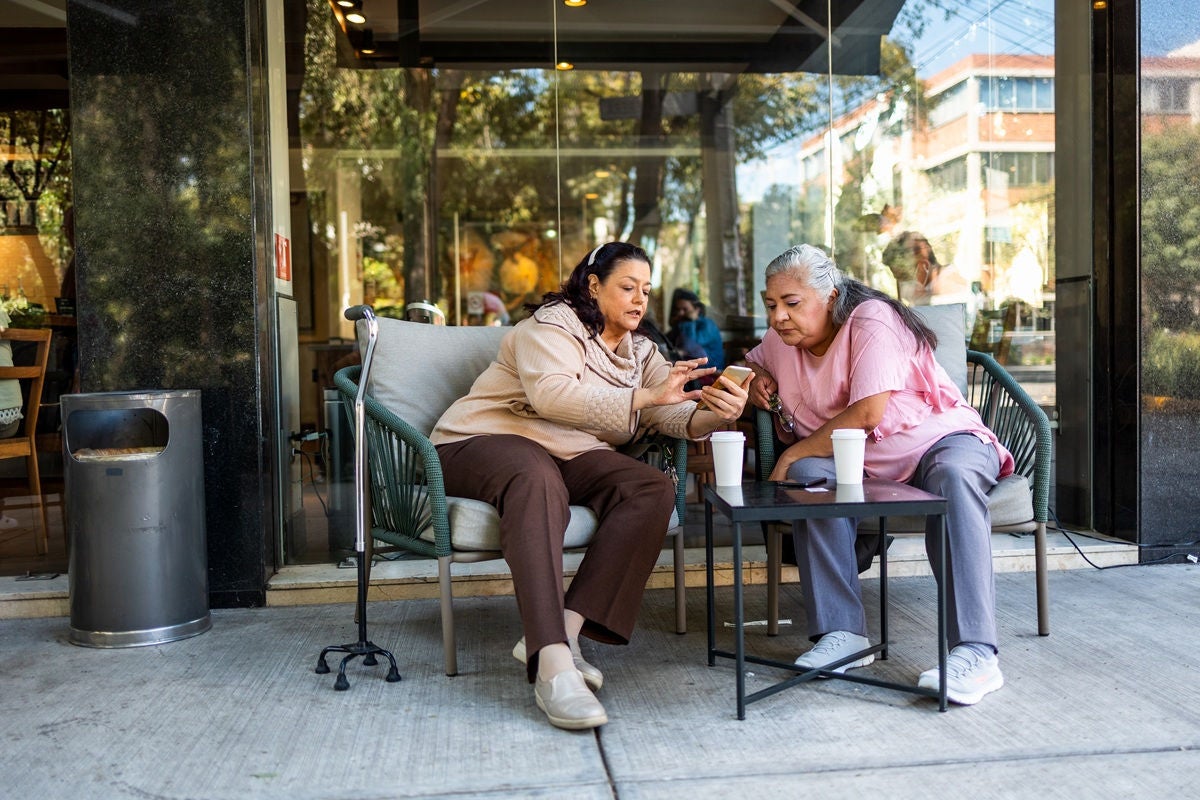 Two people seated at an outdoor café, looking at a smartphone together across a small table with coffee cups, with a glass storefront and street scene in the background.