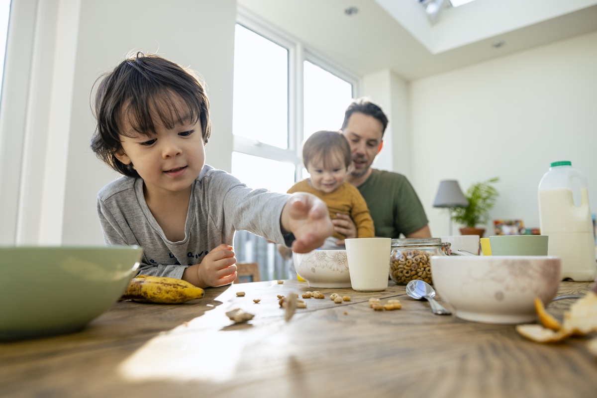 A young child reaches across a breakfast table while an adult holds a baby nearby, with bowls, cereal, fruit, and milk spread out in a sunlit kitchen.