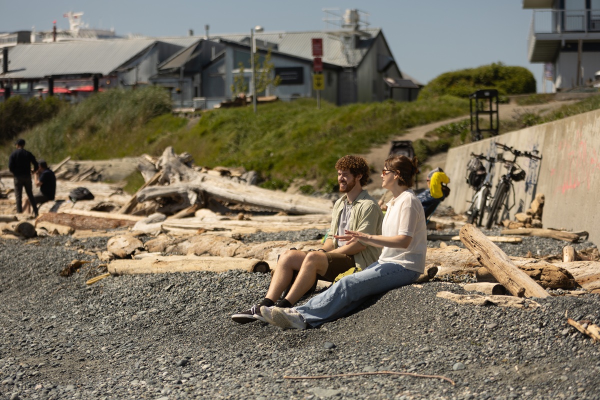 Two people sit on a rocky beach among driftwood, talking and enjoying the sunshine near a waterfront with buildings and grassy slopes in the background.